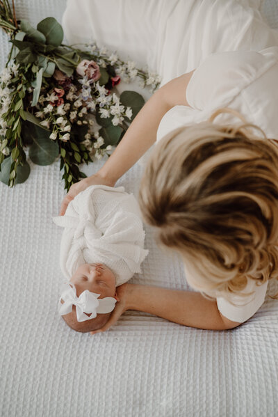 Photo of newborn baby girl wrapped in white blanket on bed with flowers next to mom and baby in Annapolis Maryland.
