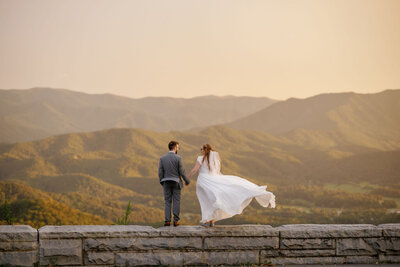Gatlinburg photographers captures couple on the stone wall in the Foothills Parkway with the wind blowing the brides gown as she holds the grooms hand and smiles to him with the Smoky Mountains in the distance while they elope to Gatlinburg 