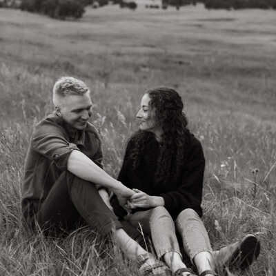 The couple sit side by side in a field for their summertime engagement session in Colorado. The field at the base of the mountains in Evergreen was the most beautiful shade of green.