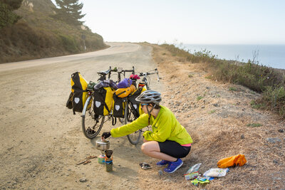 Becky makes coffee on a jetboil while two bikes balance against each other behind her during a bike tour.