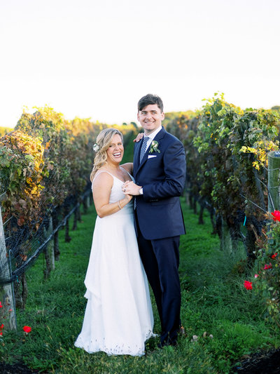 Groom wearing navy blue suit from Charles Tyrwhitt and bride wearing dress from BHLDN hold hands as the sun is setting in the vineyard at RGNY after their wedding ceremony.