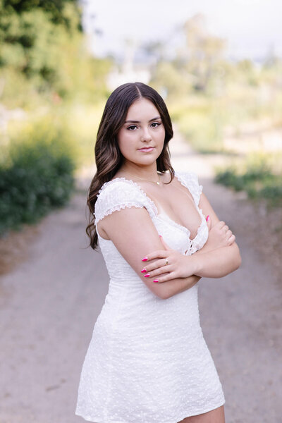 A brown haired young woman wearing a white dress leaning against a wooden gate while looking at the camera