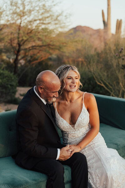 Newly married couple sitting on an emerald green sofa by the pool embracing and giggling with each other during sunset