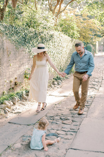 Mother and father holding hands and walking towards baby sitting on the cobblestone street in Charleston beside a brick and ivy wall