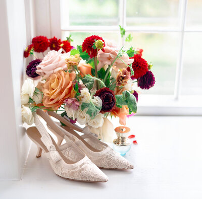 A pair of elegant Dior lace heels are placed on a windowsill next to a colorful bouquet of roses and other flowers, with a small Versace perfume bottle nearby. Bright natural light enters through the window.