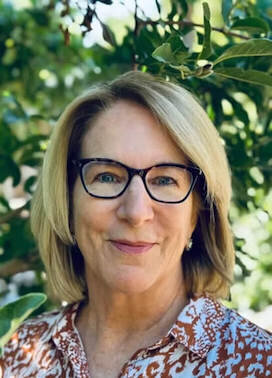 Mature woman wearing glasses smiling in an outdoor setting with green leaves in the background.