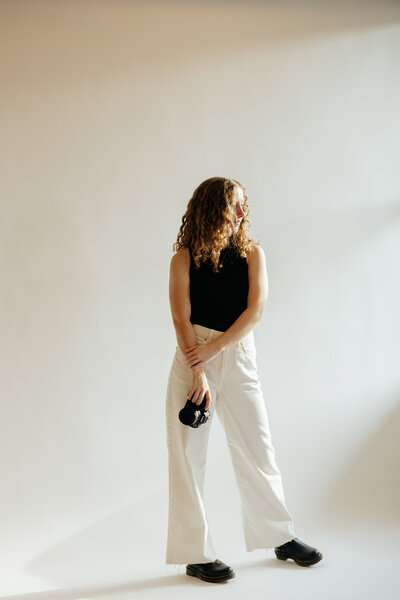 Autumn the wedding photographer standing in front of an off white back drop, looking away and holding her camera.