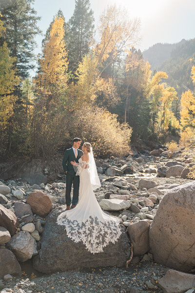 A bride and groom smile at each other while holding hands and walking towards the camera carrying skis