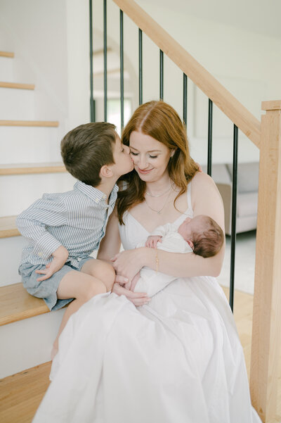 Mom sits on her staircase leaning against the rails and holding her baby while her toddler sits next to her and kisses her cheek