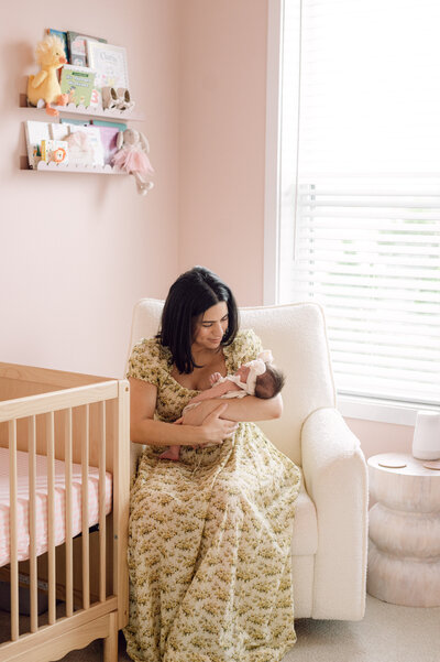 A mother smiles while holding her newborn baby in a cozy pink nursery, captured by a Northern Virginia newborn and family photographer during an in-home session.