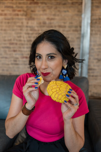 The founder of Soleil Vida Studio as she discusses art workshops, seated on a sofa and enjoying a typical Mexican dish. Adorned in a vibrant fuscia blouse and long blue earrings, this intimate moment captures the essence of creativity and cultural celebration