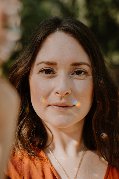 The Intuitive Momma looks into camera and smiles softly as rainbow light fractals reflect on her face. The Intuitive Momma offers a variety of services, including 1:1, event or retreat reiki and somatic sessions.