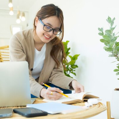 WOMAN WITH BROWN HAIR AND GLASSES WEARING BEIGE CARDIGAN AND JEANS WRITING IN A NOTEBOOK WITH LAPTOP ON TABLE