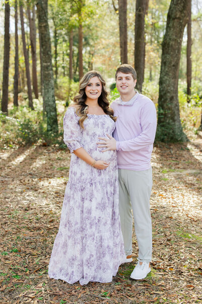 mom and dad standing in trees holding moms pregnant belly