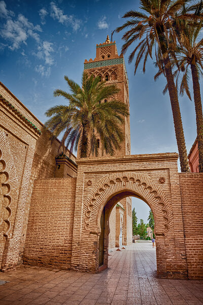 A tall, ornate brick tower stands under a blue sky, framed by slender palm trees. The intricate archway in front features detailed brickwork, leading to a paved pathway. The architecture has a historical and cultural touch.