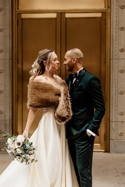 Bride in elegant gown and fur wrapped around shoulders holds bouquet and looks lovingly at husband in tux.
