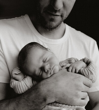 Black and white photo of a father holding their newborn.  The photo is cropped so you cannot see the father's face fully, the baby is close to his chest facing outwards and sleeping.  