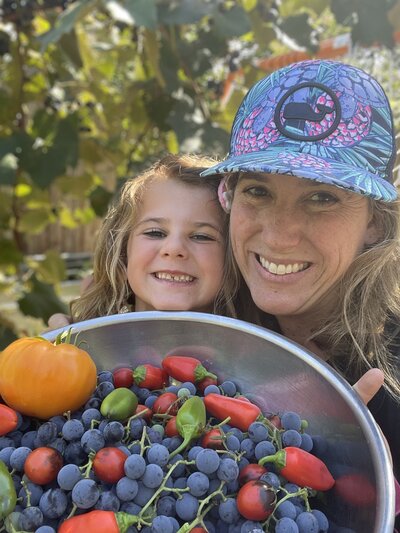 A smiling woman and a child holding a bowl full of fresh garden produce, including grapes, tomatoes, and peppers, with green foliage in the background.