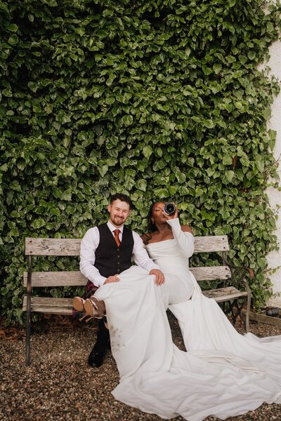 A couple sit side by side on a bench and drink prosecco from the bottle. They wear a dress and a kilt and pose for photographs.