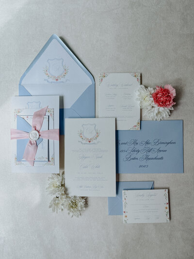 White Velvet ring box containing 3 rings, a men's watch, with greenery and white flowers, and wedding invitation suite in the background.