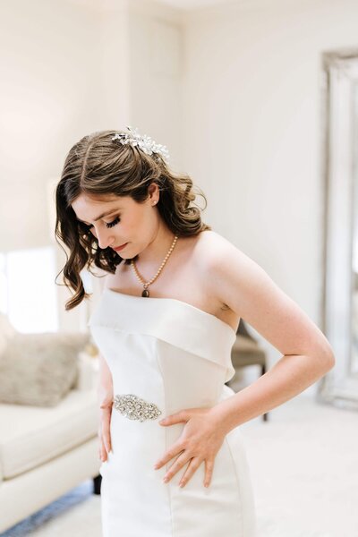 A bride with wavy brown hair and a hairpiece looks down as she adjusts her strapless white wedding dress, standing in a softly lit room with a sofa and a large mirror in the background.