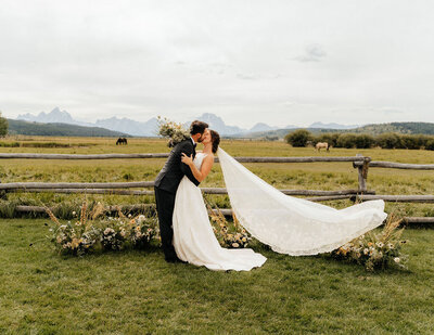 bride and groom kissing in a field next to a wooden fence with mountains in the background