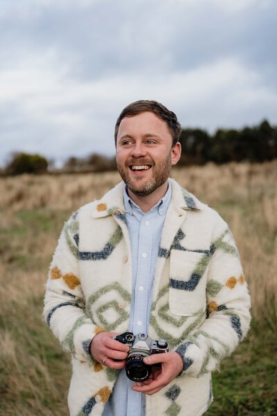 Aberdeen wedding photographer Scott Arlow stands in a large field and holds a camera. He wears a blue shirt and a colourful jacket.