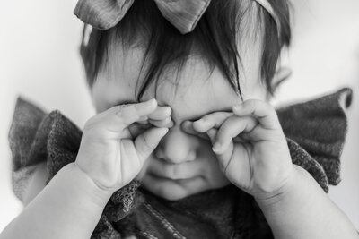 A delicate black and white close-up of an infant’s tiny fingers rubbing her sleepy eyes, capturing a quiet, fleeting moment of early childhood.