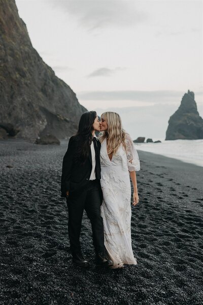 LGBTQ+ couple kissing on Reynisfjara Black Sand Beach in Iceland during their intimate elopement photographed by Destination Elopement Photographer, with dramatic cliffs and sea stacks in the background.