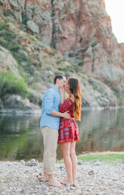 Engaged couple share a a kiss in front of desert mountain. They stand on rocks by a stream of water.