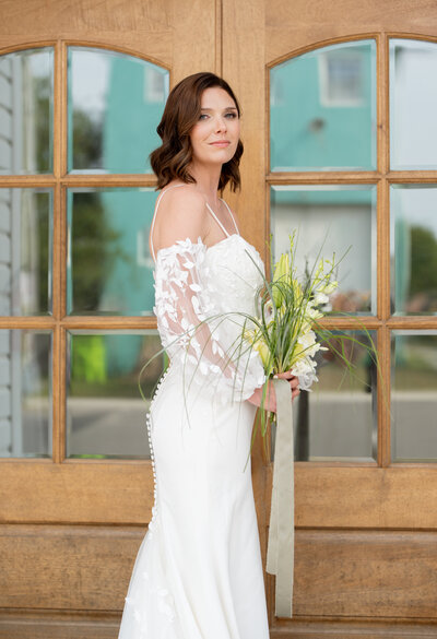 A picture of a bride getting her portrait holding a white and green bouquet. Standing in front of double doors. 