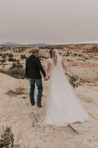 The groom who is wearing a tan cowboy hat is holding hands with his bride. Their backs and to the camera and they are walking away from the camera into the wild landscape of Capitol Reef National Park.