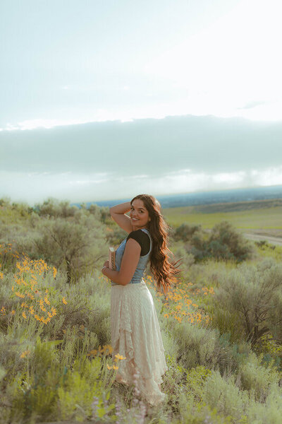 A woman stands on a hill with sagebrush and other greenery. She's looking over shoulder smiling at the camera.