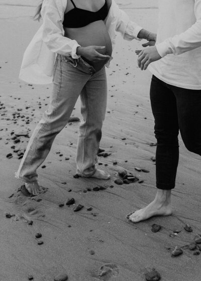 a black and white images of a couple on the beach in florida in loose denim, barefoot, and showing the womans maternity belly