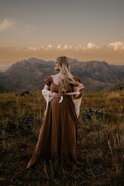A pregnant woman stands in the mountains holding her baby belly