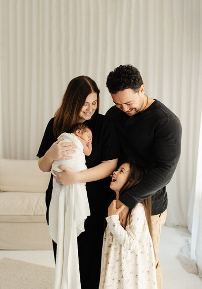A family of four looking at one another and laughing while the mum holds the newborn baby and lucy takes candid photographs in her christchurch newborn photo studio