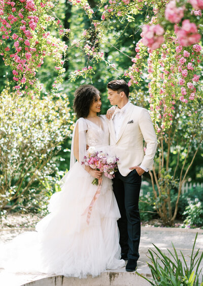 bride and groom surrounded by pink roses in a garden smiling by Colorado Wedding Photographer JKG Photography