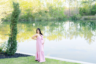 Woman standing by water in a pink dress holding her pregnant belly smiling at the camera for maternity photography