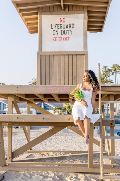 A brunette young woman in a white dress sitting on a lifeguard tower in Newport Beach