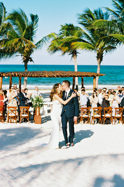 Bride and groom smiling as they exchange vows at the Liberty Warehouse in Brooklyn New York, under a floral arch by the ocean, surrounded by sunlight and laughter — destination wedding documented by Asia Pimentel Photography.