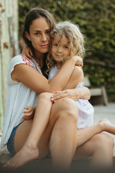 A mother and toddler cuddling a in a beautiful motherhood moment , they are outdoors and looking a the camera 