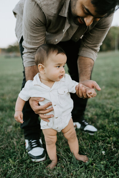 A baby stands in the grass with the help of his dad holding his hands in Lafayette, Indiana.