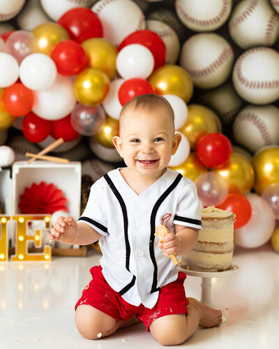 A baby boy kneels near a cake, holding a spoon and smiling, in front of a baseball background
