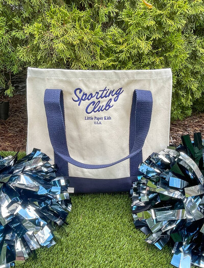 Canvas tote bag with navy blue straps and embroidered Sporting Club logo sitting next to two pom poms