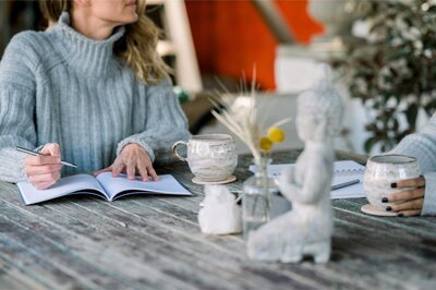 Two women sit at a rustic wooden table with journals and ceramic mugs, surrounded by soft natural decor and a small Buddha statue.