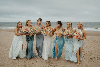 Bride and her bridal party that are dressed in different shades of blue are carrying colorfully vibrant bouquets and laughing with their arms linked and walking on the beach