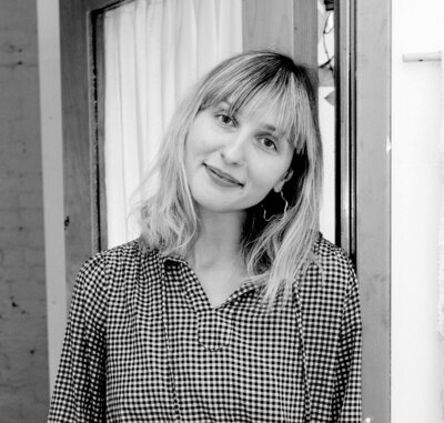 Black and white photo of woman with short blonde hair and bangs softly smiles at the camera while leaning against a doorframe.