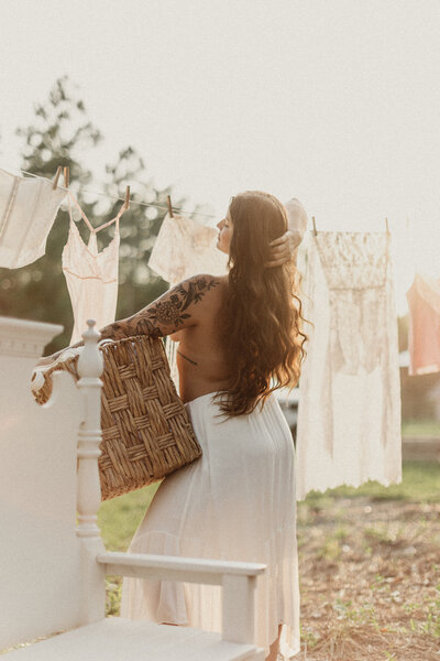 Woman with long red hair and tattooed arm stands topless in front of vintage laundry line, lit by golden hour light during Jacksonville boudoir session.