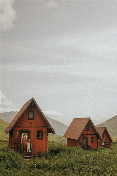 A couple stands on the porch of a small, rustic red cabin in a remote, grassy area in Alaska. The cabin is nestled in a valley with distant mountains under a cloudy sky.