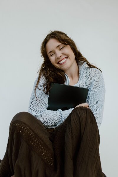 A website designer holding her laptop while sitting on the ground in front of a white backdrop.